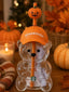 A Starbucks bear-shaped clear glass cup holding an orange Starbucks straw with a pumpkin topper and an orange cap printed with the Starbucks logo, styled on a wooden surface with pumpkins, autumn leaves, a candle, and a blanket in the background.
