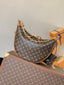 A Louis Vuitton crescent-shaped shoulder bag in brown monogram canvas with gold-tone chain strap, displayed on top of a matching monogram trunk with orange Louis Vuitton boxes in the background.