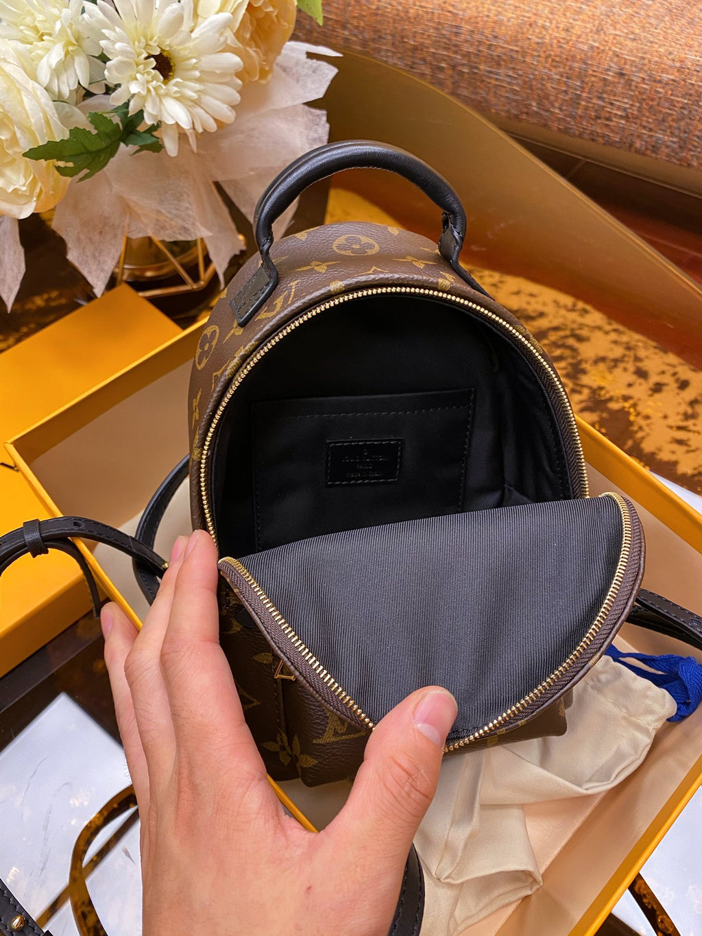 A close-up of a hand holding open a small brown monogram backpack, showing the black interior lining and zipper. The backpack is placed inside a yellow Louis Vuitton box, with a bouquet of white flowers in the background.