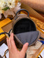 A close-up of a hand holding open a small brown monogram backpack, showing the black interior lining and zipper. The backpack is placed inside a yellow Louis Vuitton box, with a bouquet of white flowers in the background.