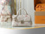 A Louis Vuitton handbag in white textured leather with the brand's classic multicolor monogram pattern, featuring silver-tone chain straps and a turn-lock clasp, displayed on a marble table with orange Louis Vuitton packaging in the background.