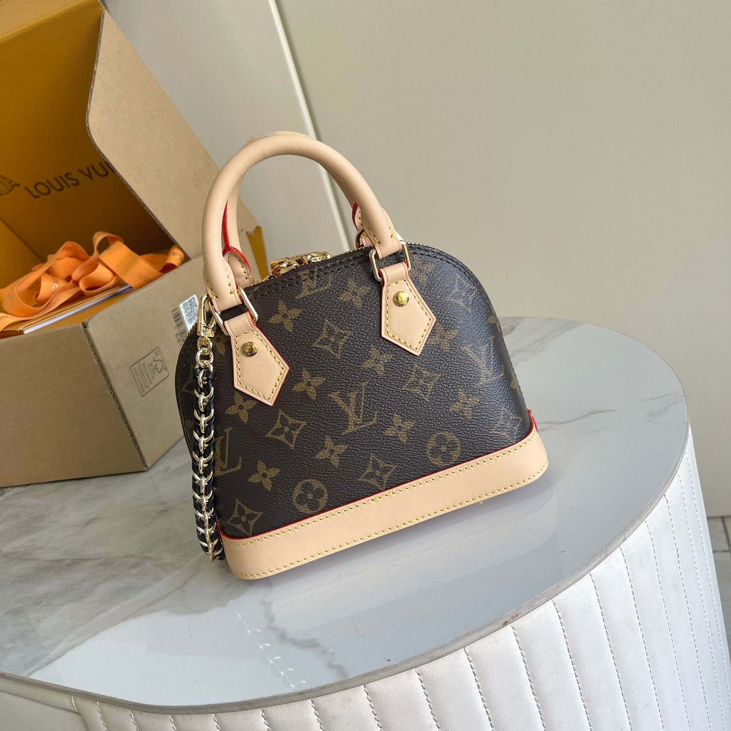 Louis Vuitton mini Alma handbag in classic monogram canvas with beige leather trim and handles, displayed on a white marble-topped table next to its branded packaging box.