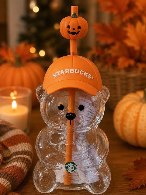 A Starbucks bear-shaped clear glass cup holding an orange Starbucks straw with a pumpkin topper and an orange cap printed with the Starbucks logo, styled on a wooden surface with pumpkins, autumn leaves, a candle, and a blanket in the background. Main image