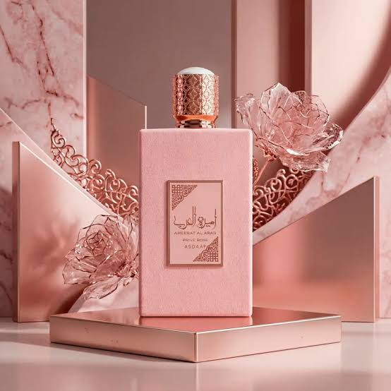 A pink rectangular perfume bottle with a rose-gold cap and a front label reading ASADANA, styled on a metallic platform with pink marble and rose-shaped decorative props.