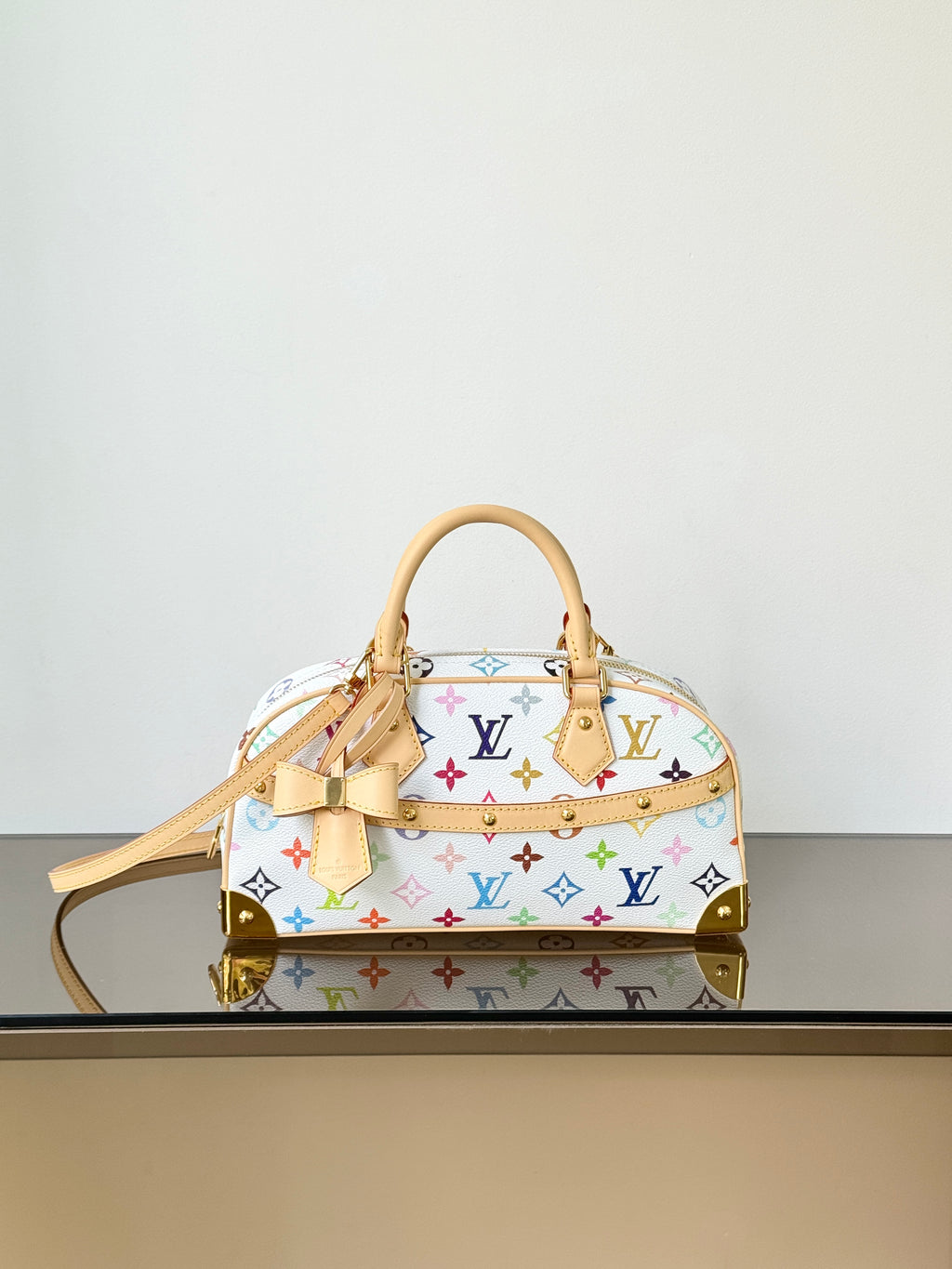 A Louis Vuitton monogram handbag in white coated canvas with multicolor printed monogram motifs, tan leather handles and trim, and gold-tone hardware, displayed on a reflective surface against a plain light background.