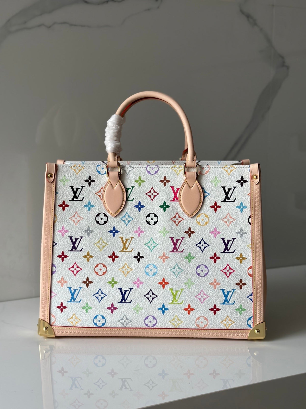 A Louis Vuitton handbag with a white canvas body covered in a colorful multicolor monogram pattern, featuring light pink leather trim, double top handles, and gold-tone hardware, set against a gray marble background.