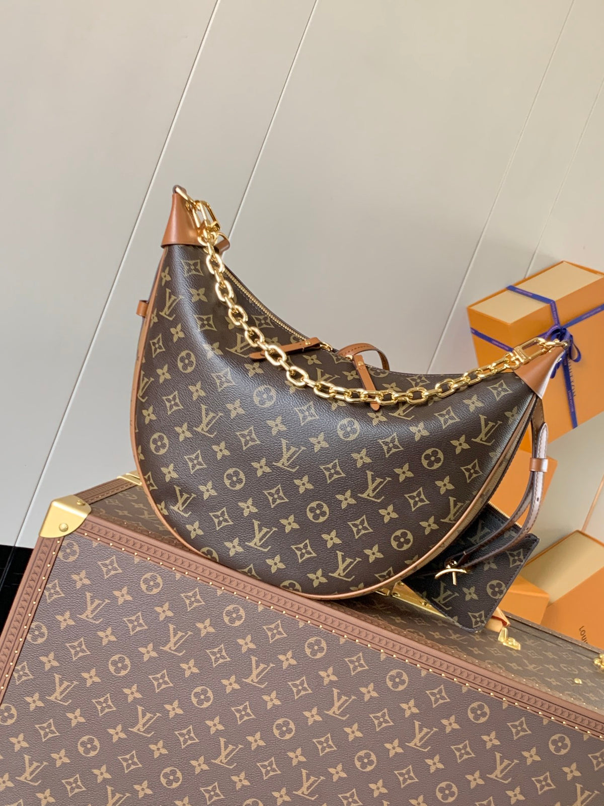 A Louis Vuitton crescent-shaped shoulder bag in brown monogram canvas with gold-tone chain strap, displayed on top of a matching monogram trunk with orange Louis Vuitton boxes in the background.