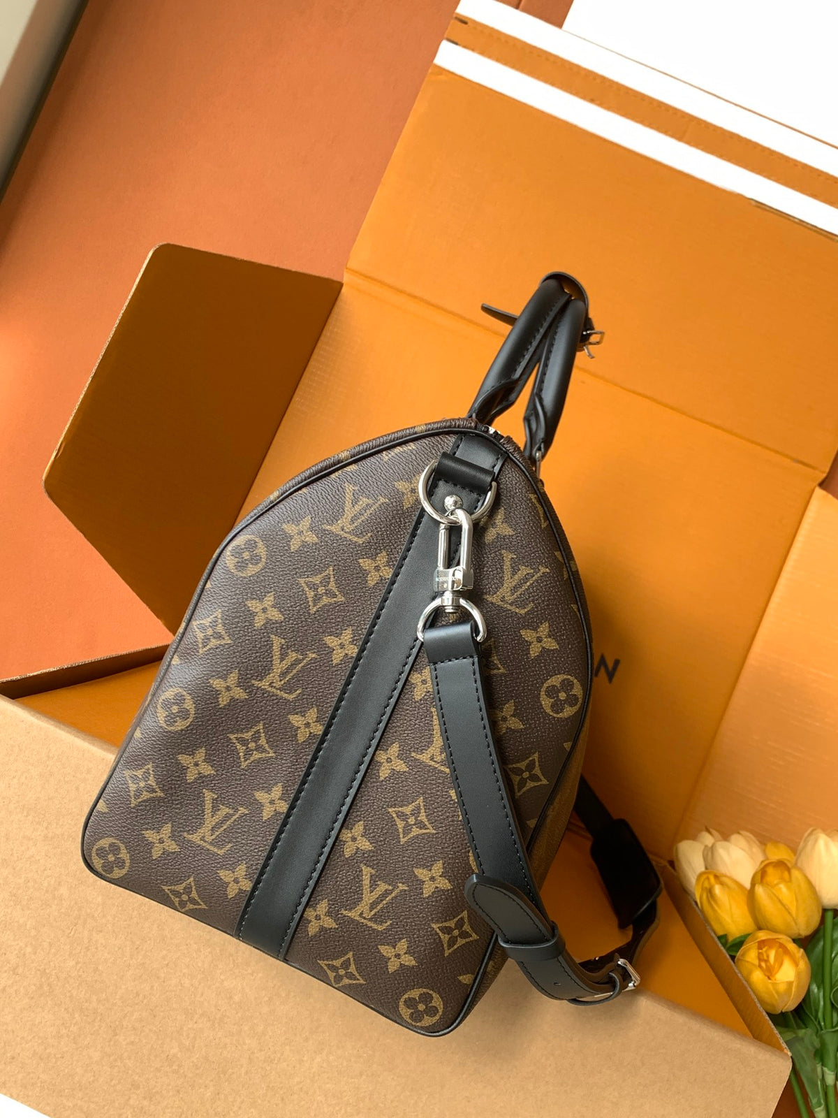 A Louis Vuitton monogram shoulder bag in brown coated canvas with black leather trim and strap, displayed on an orange Louis Vuitton box with yellow tulips in the corner.