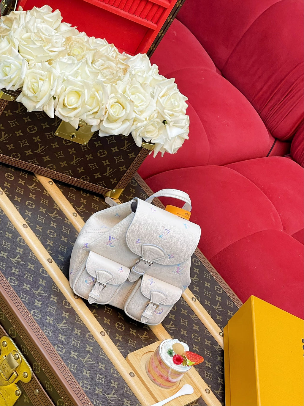 A small white backpack with a subtle multicolor monogram pattern sits on a Louis Vuitton suitcase next to a red velvet sofa and white roses.